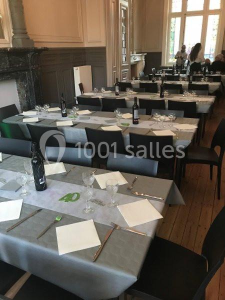 Salle à manger avec tables dressées, nappes grises, couverts, verres et bouteilles de vin, éclairée par des fenêtres.