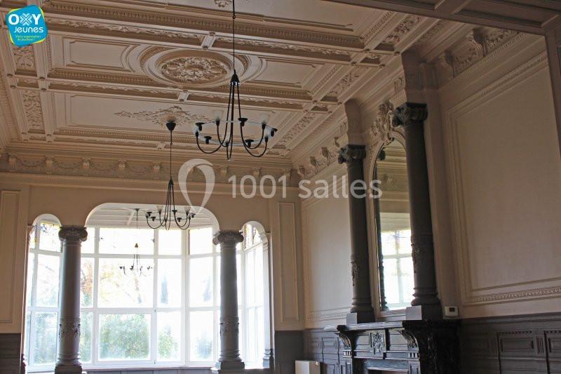 Plafond orné et mouluré d'une salle lumineuse avec grandes fenêtres, colonnes et cheminée en bois sculpté.