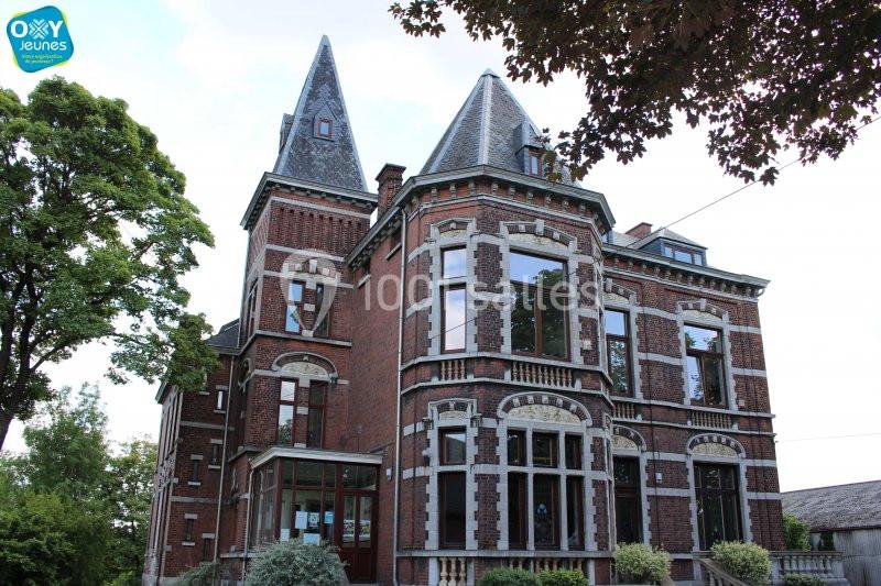Grande maison en briques rouges avec deux tours, entourée d'arbres et d'arbustes, sous un ciel dégagé.