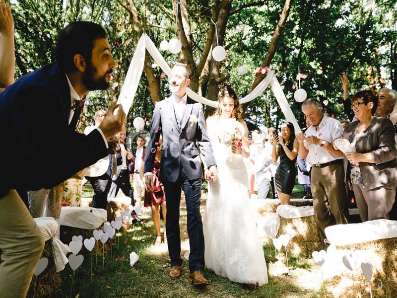 Un couple souriant marche dans une allée décorée, entouré d'invités célébrant en plein air sous des arbres.