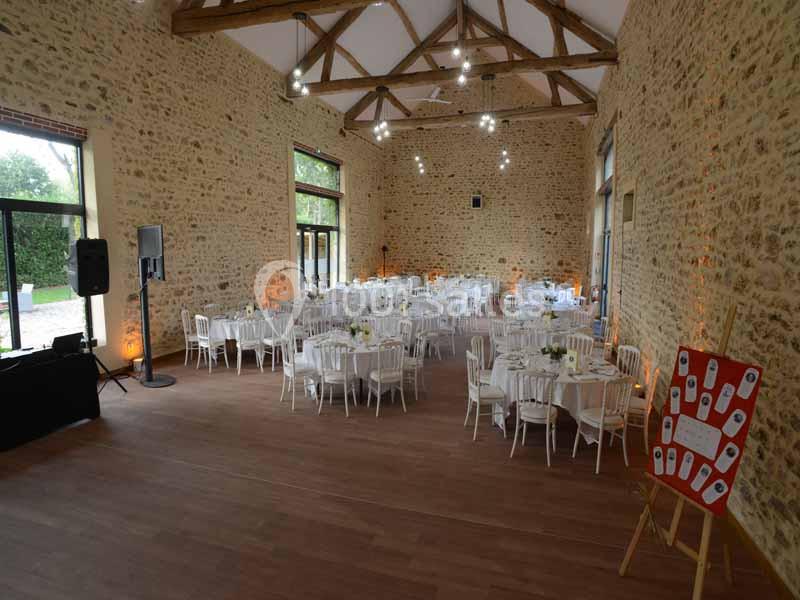 Salle de réception en pierres apparentes avec tables rondes dressées, éclairée par des suspensions et des baies vitrées.