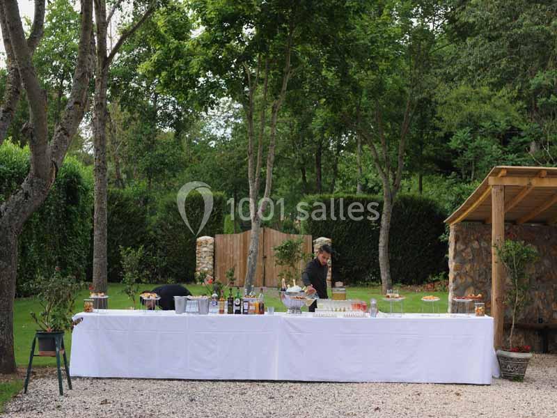 Table dressée en extérieur avec nappes blanches, boissons et plats, entourée d'arbres et d'un jardin verdoyant.