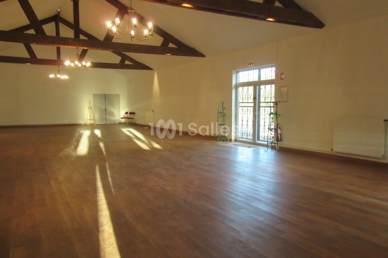 Salle spacieuse avec parquet en bois, poutres apparentes, grandes fenêtres et lumière naturelle entrant.