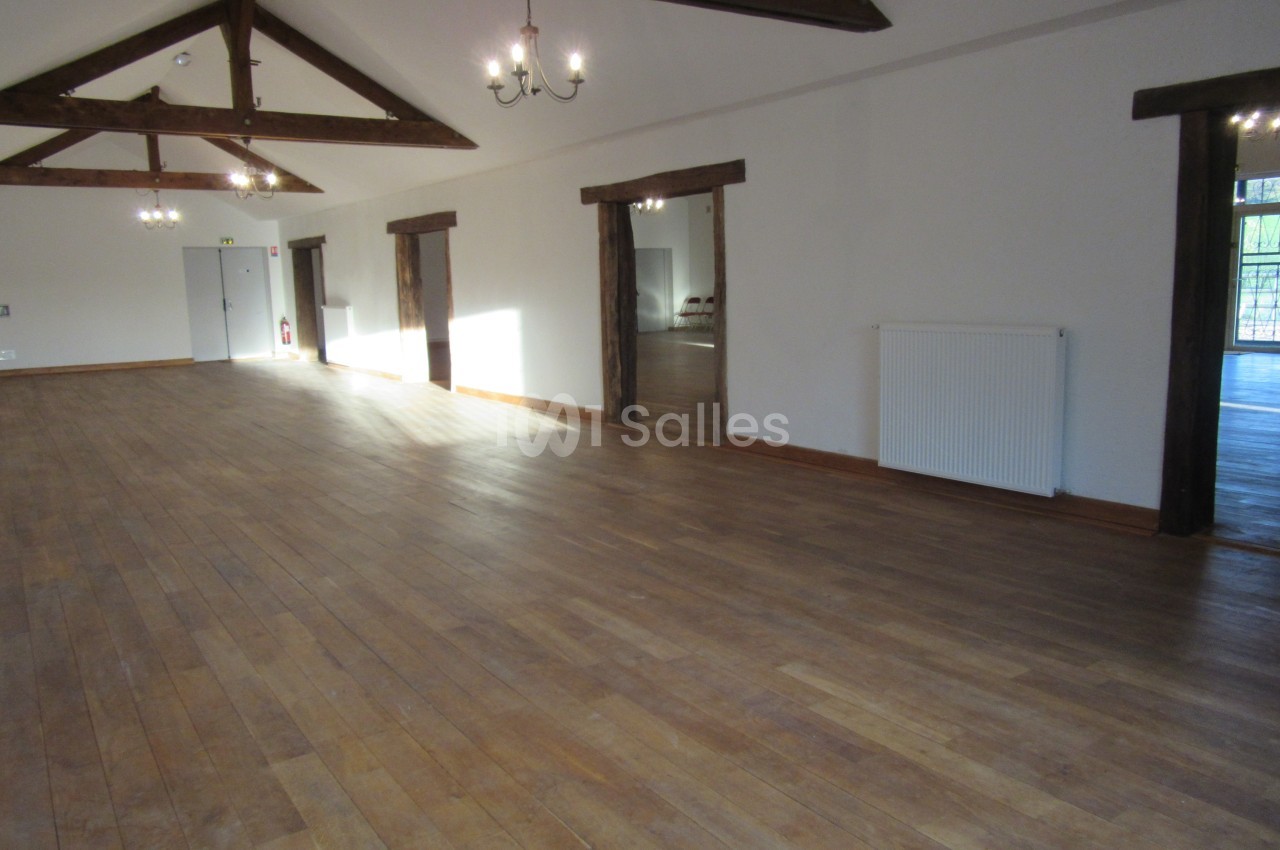 Salle spacieuse avec parquet en bois, poutres apparentes au plafond et lumière naturelle entrant par des fenêtres.
