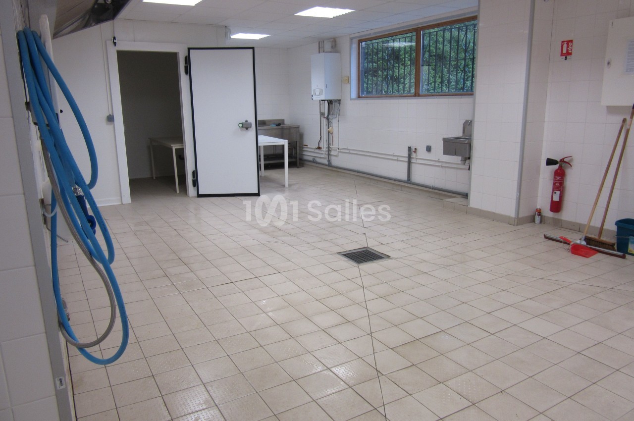 Salle carrelée vide avec murs blancs, éclairage au plafond, porte ouverte, tuyaux apparents et équipements de nettoyage.