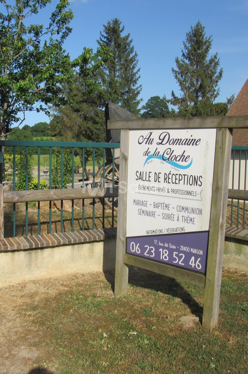 Panneau en bois indiquant ’Au Domaine de la Cloche’, salle de réceptions, devant une clôture et un paysage rural.