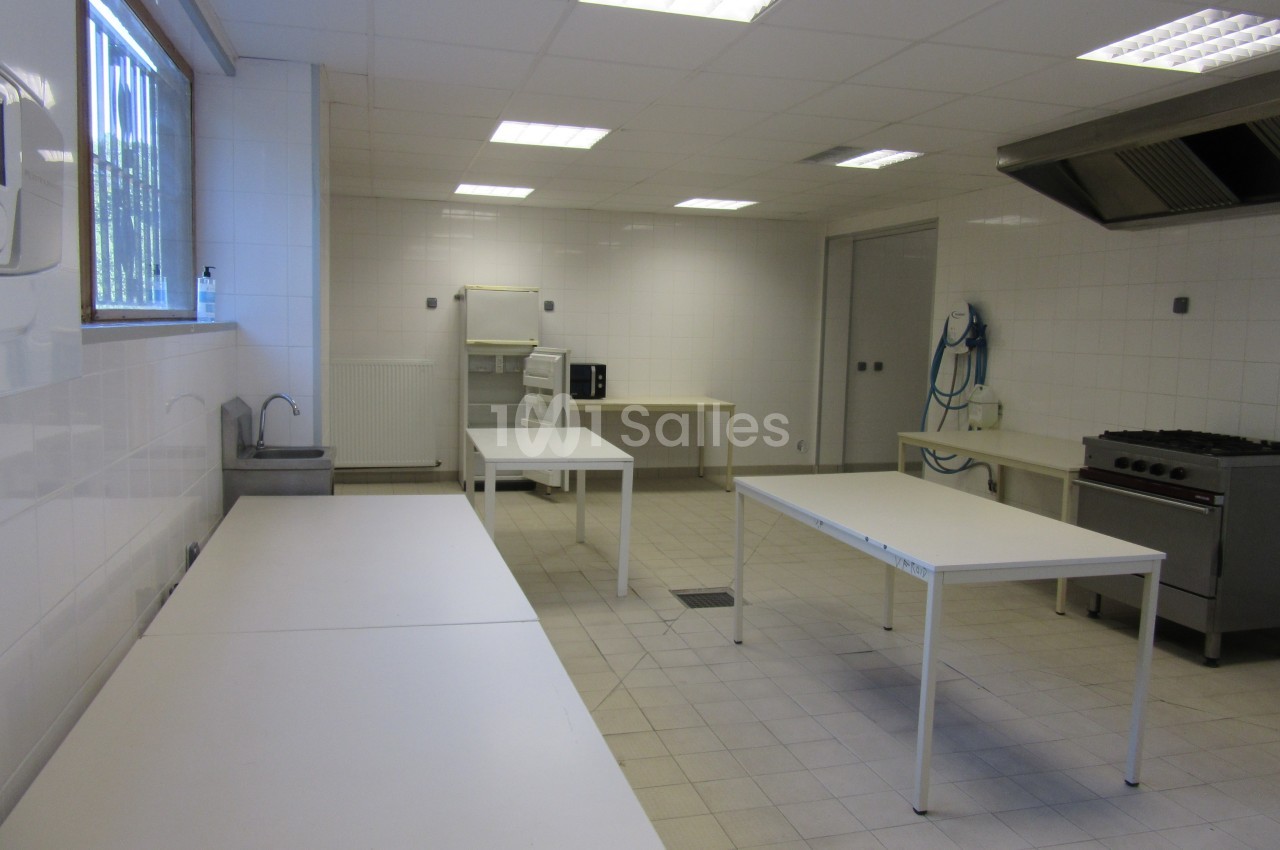 Cuisine professionnelle vide avec plans de travail blancs, évier, hotte, four et carrelage clair.