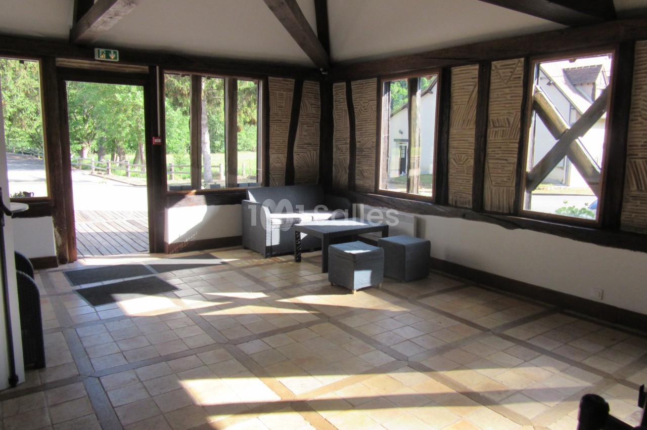 Salle lumineuse avec grandes fenêtres, mobilier en rotin gris et poutres apparentes, donnant sur un espace extérieur…