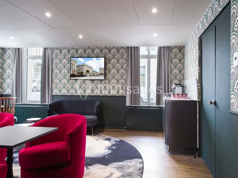 Salle lumineuse avec fauteuils rouges, table noire, tapis rond, télévision murale et coin boissons sur un meuble.