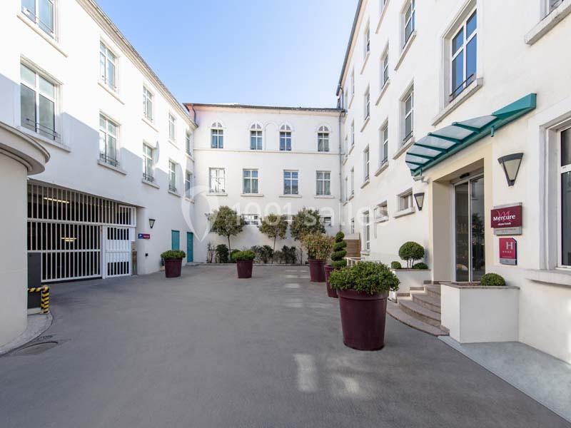 Cour intérieure d'un bâtiment blanc avec des fenêtres, des plantes en pots et une entrée d'hôtel sur la droite.