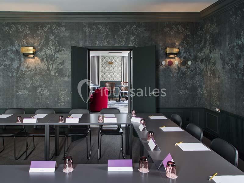 Salle de réunion élégante avec tables disposées en U, chaises noires et vue sur un salon décoré de fauteuils rouges.