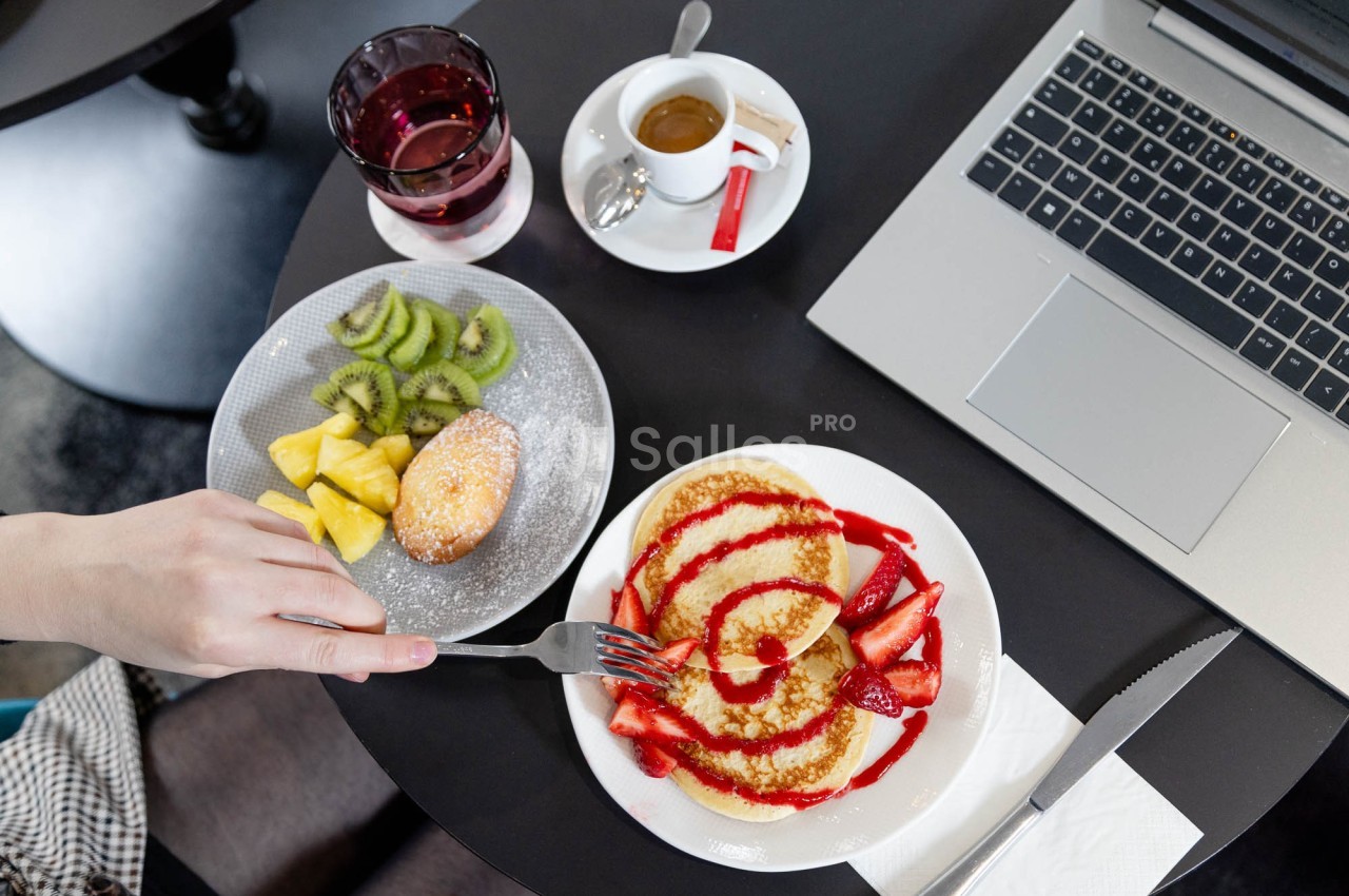 Assiette de pancakes garnis de fraises et coulis, accompagnée de fruits, café et ordinateur sur une table noire.