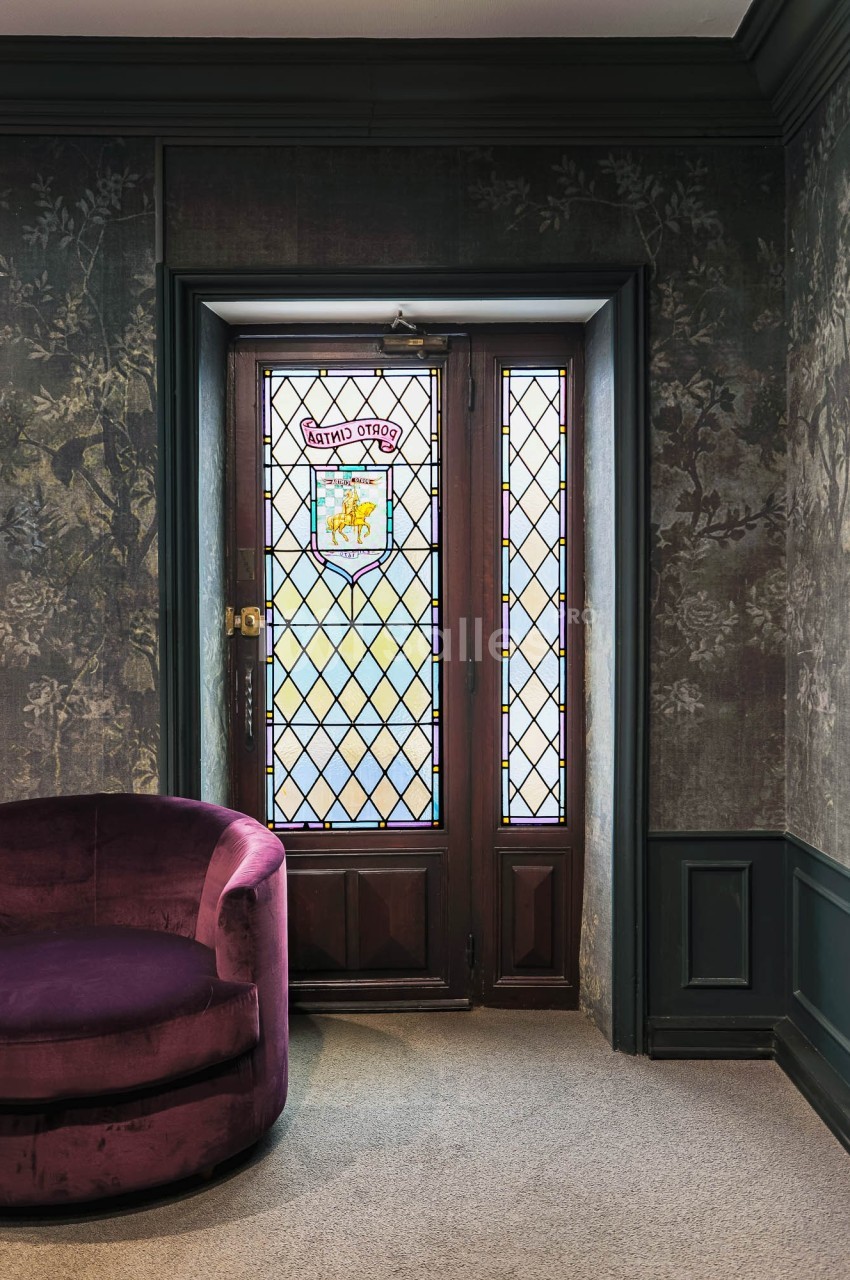 Porte en bois avec vitraux colorés, encadrée par un mur sombre et un fauteuil violet au premier plan.