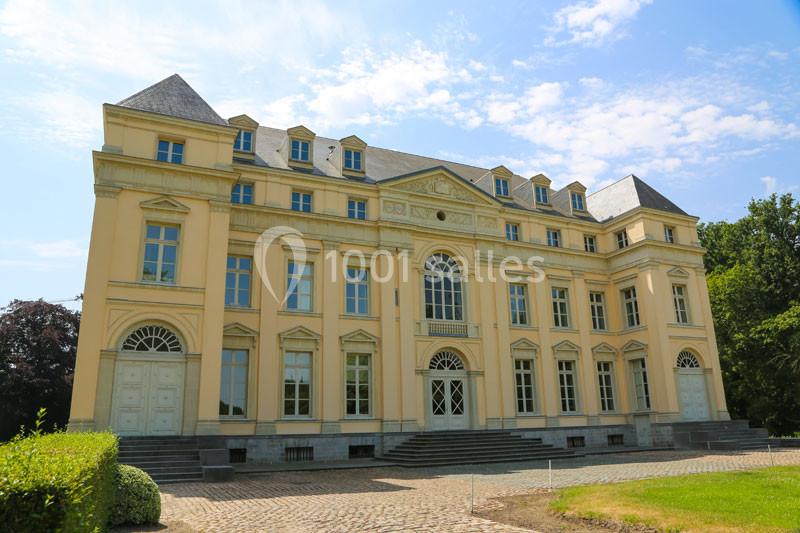 Façade d'un grand bâtiment classique beige avec des fenêtres symétriques, entouré de verdure et d'un ciel dégagé.