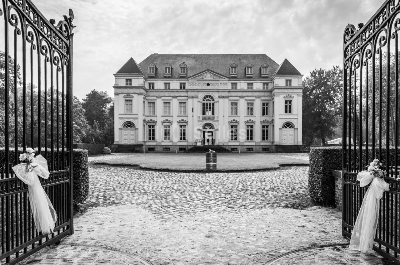 Entrée principale d'un château entourée de grilles ouvertes, avec des décorations florales et une allée pavée.