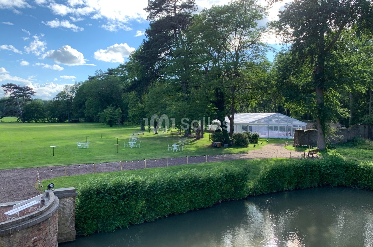 Vue d'un parc verdoyant avec une grande tente blanche, des arbres, des tables de jardin et une rivière au premier plan.