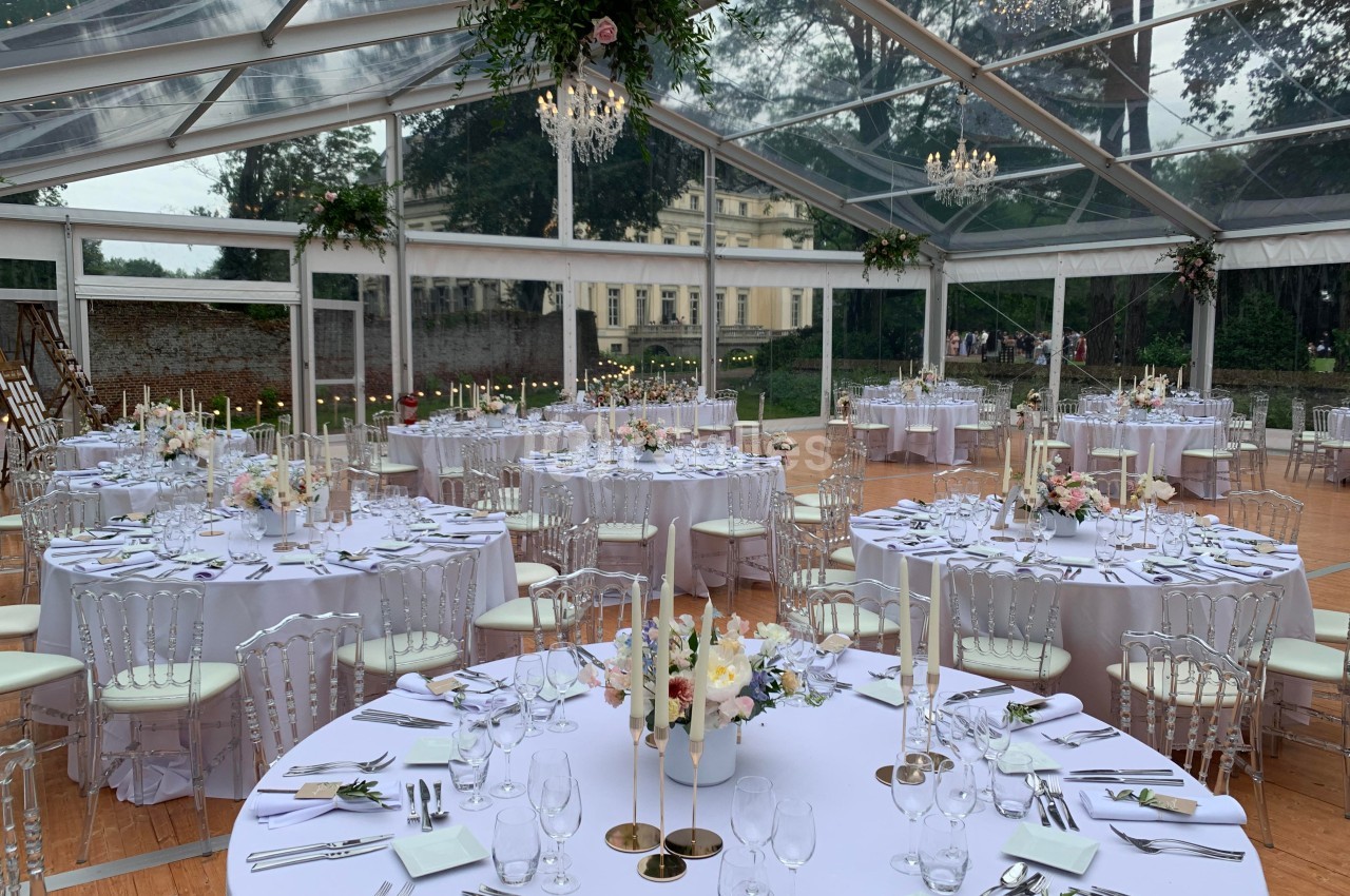 Salle de réception sous une tente transparente, tables rondes dressées avec nappes blanches et décorations florales.