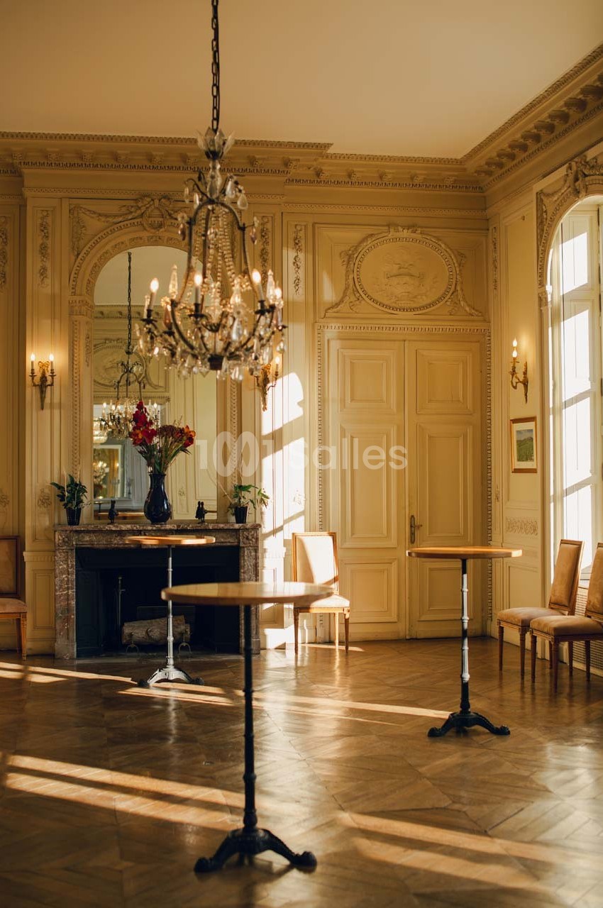 Salle élégante avec parquet en bois, tables hautes, cheminée ornée d'un miroir et lustre suspendu.