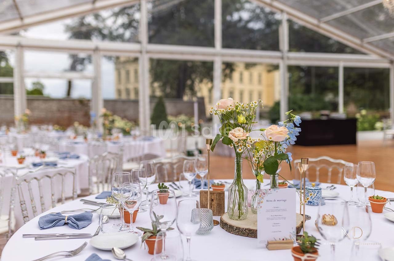 Salle de réception lumineuse avec tables décorées de fleurs, vaisselle élégante et nappes blanches sous une verrière.