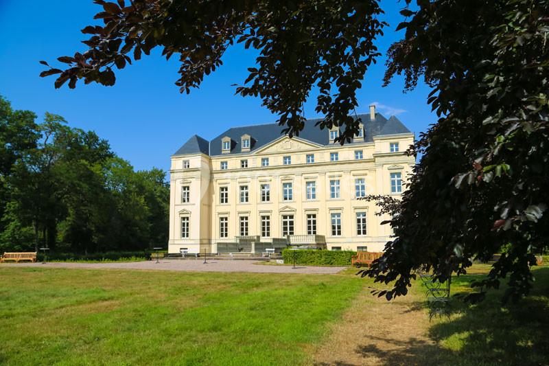 Façade d'un château classique entouré de pelouses et d'arbres sous un ciel bleu.