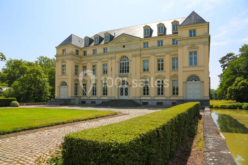 Façade d'un château jaune à trois étages, entouré de verdure, avec une allée pavée et un canal adjacent.