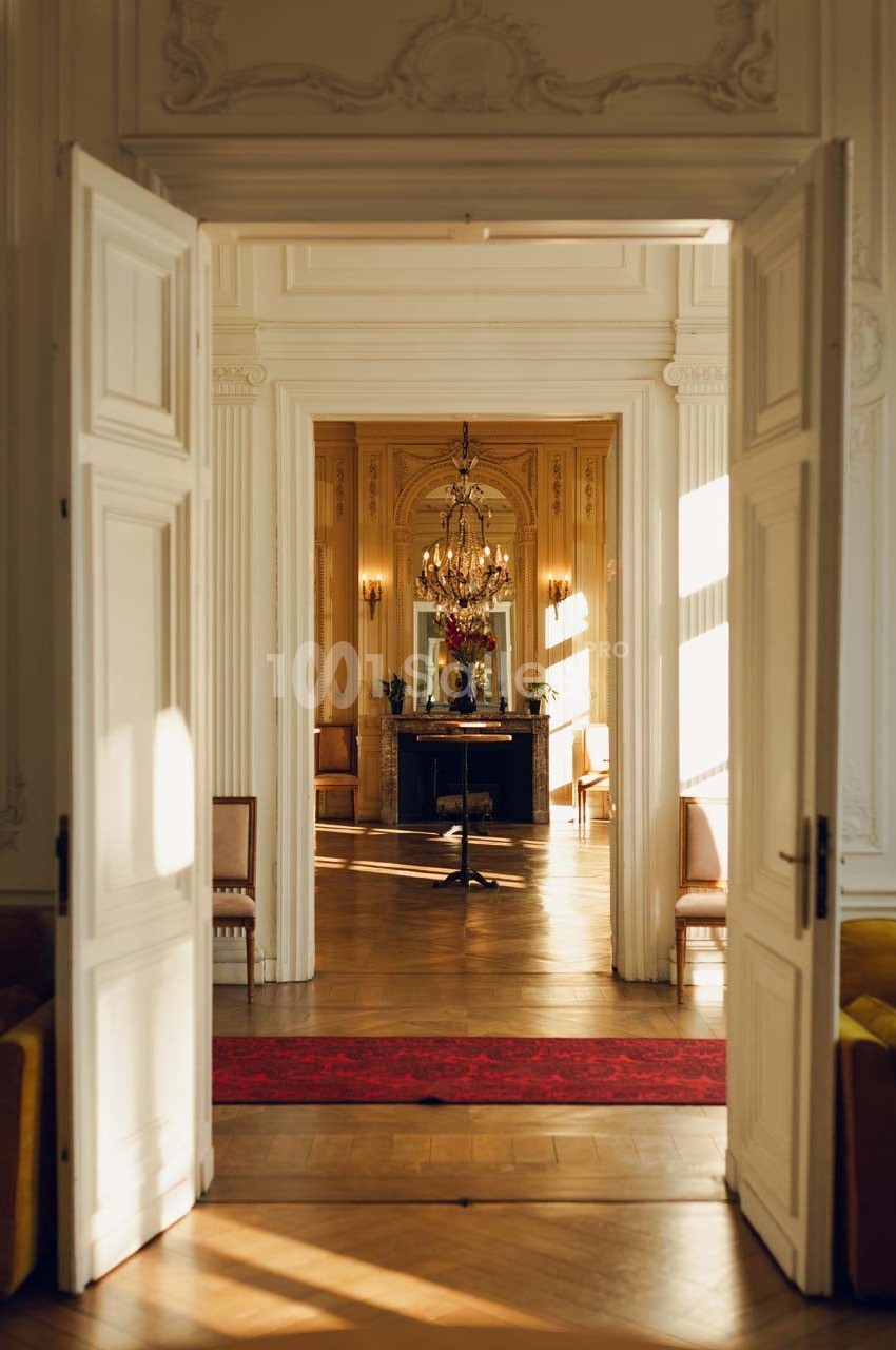 Vue d'un intérieur élégant avec des portes ouvertes, un lustre, une cheminée et un tapis rouge éclairés par la lumière…
