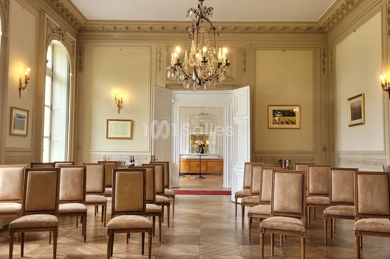 Salle lumineuse avec parquet en bois, chaises alignées, murs ornés de moulures et lustre central.