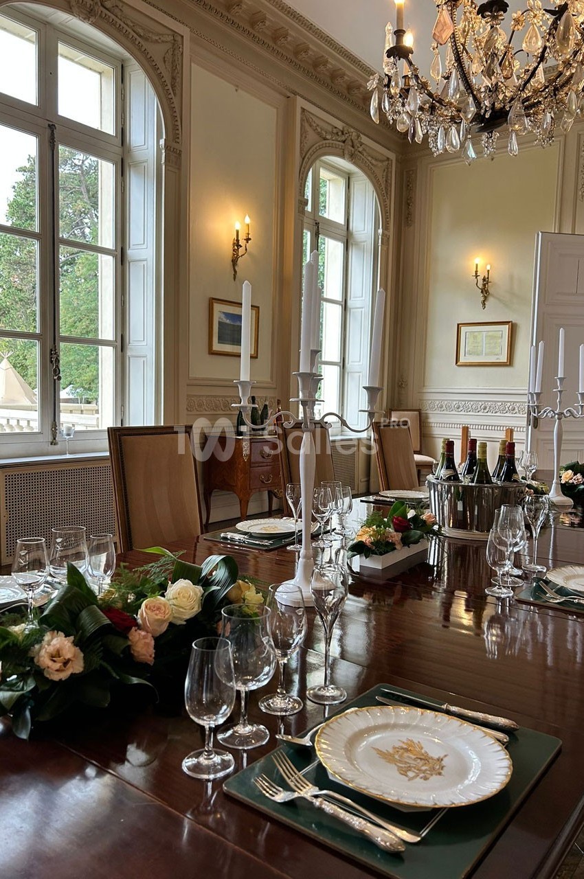 Table élégamment dressée avec vaisselle raffinée, chandeliers et décorations florales dans une salle lumineuse et classique.