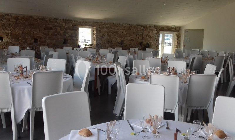 Salle de réception avec des tables rondes dressées, nappes blanches, chaises blanches et murs en pierre apparente.