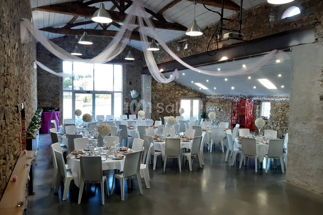 Salle de réception en pierre avec tables dressées pour un événement, décorée de voilages suspendus et éclairée naturellement.