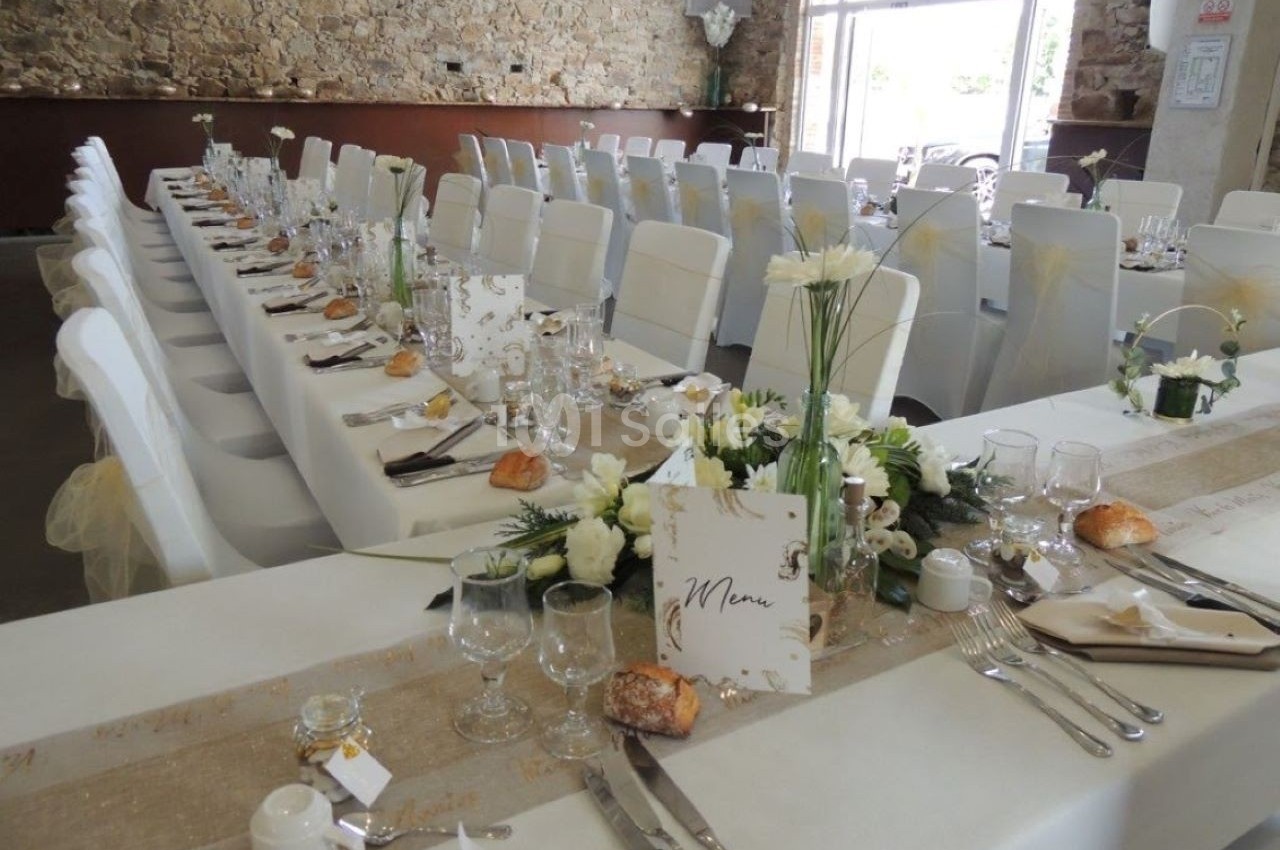 Salle de réception décorée pour un mariage avec longues tables dressées, nappes blanches et arrangements floraux.