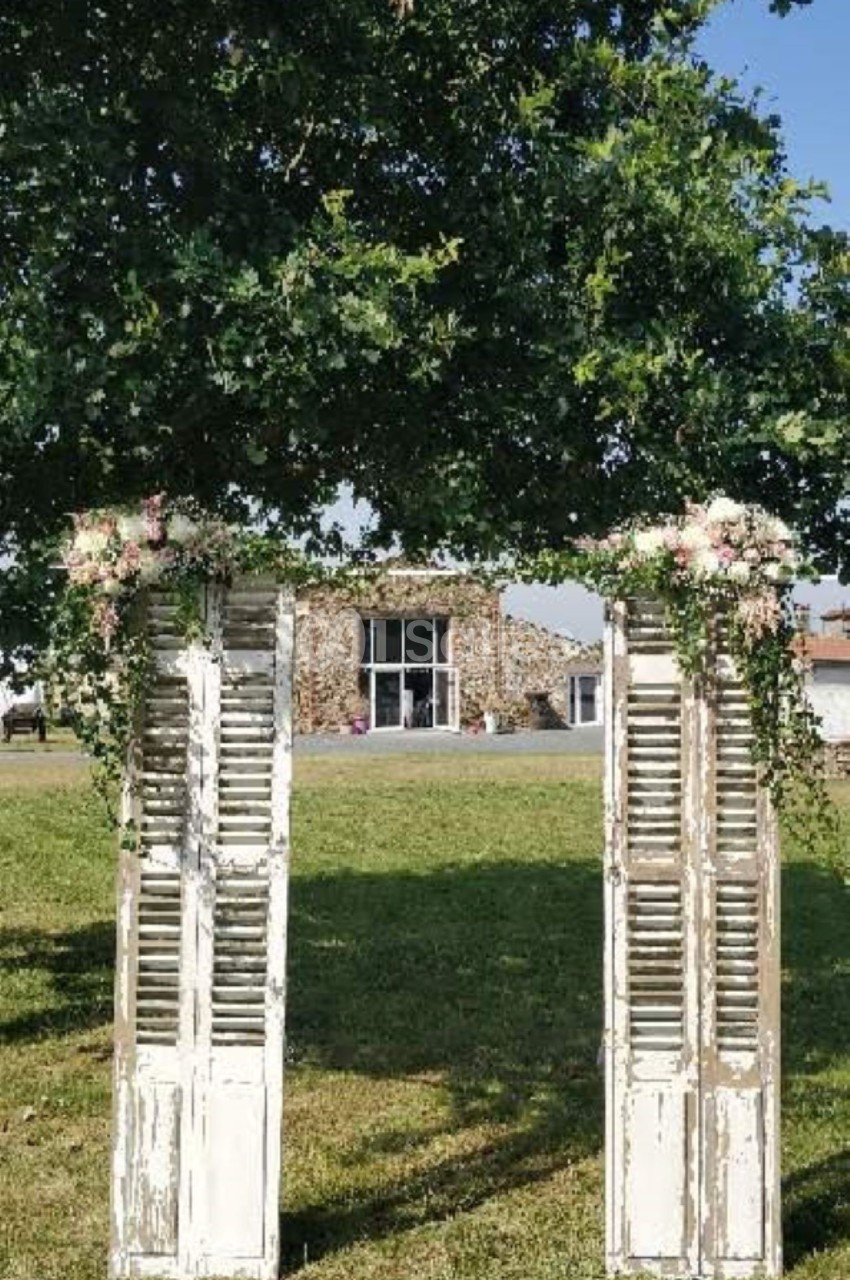 Deux volets en bois vieilli décorés de feuillage et de fleurs, formant une arche sur une pelouse devant un bâtiment en…
