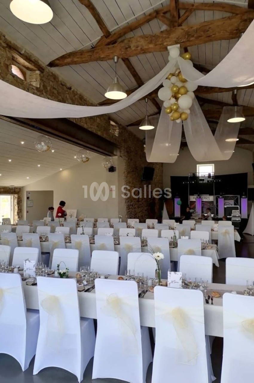 Salle de réception décorée avec des chaises blanches, des nœuds dorés, des tables dressées et des guirlandes suspendues.