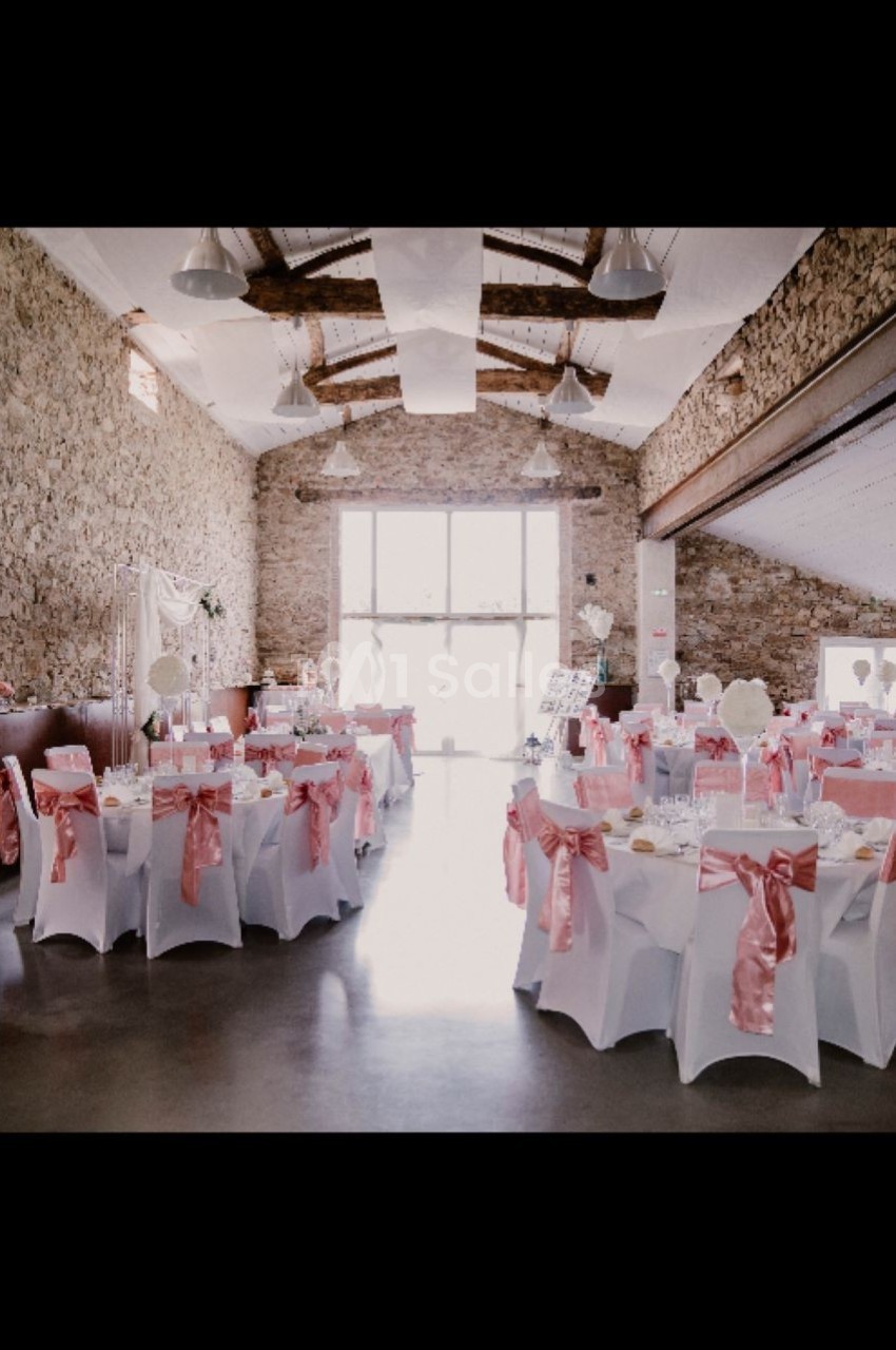 Salle de réception décorée avec des nappes blanches et des nœuds roses, éclairée par une grande baie vitrée.