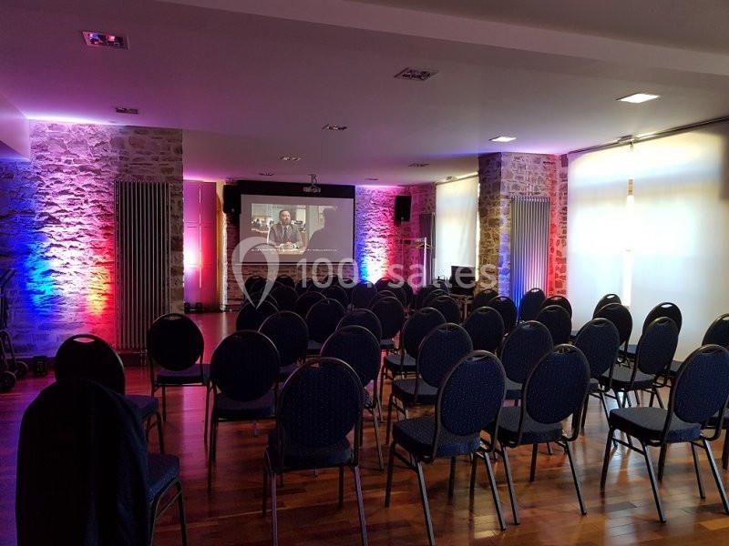 Salle de conférence avec des rangées de chaises vides, un écran de projection allumé et un éclairage coloré.