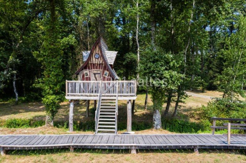 Cabane en bois sur pilotis avec toit pointu, entourée d'arbres dans une clairière, accessible par un escalier.