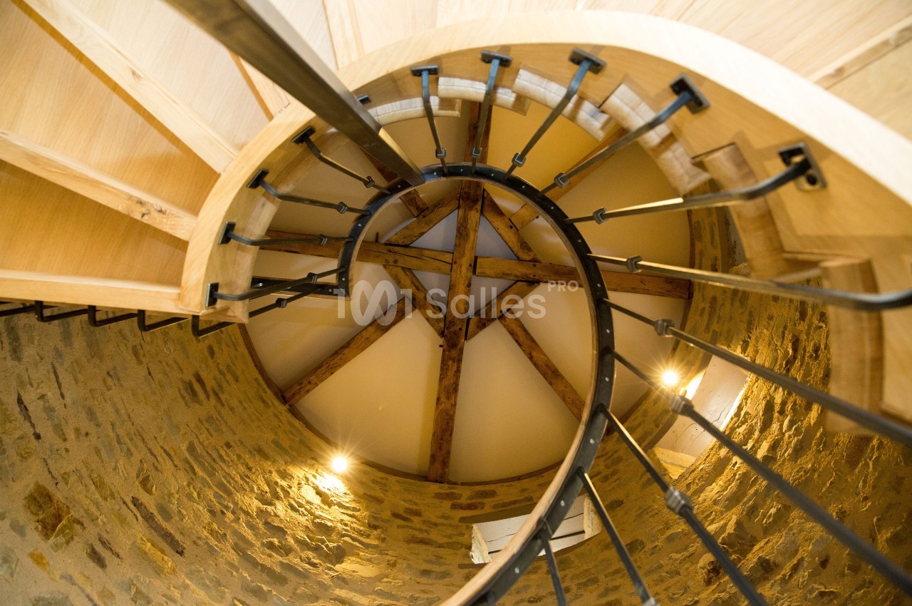 Escalier en colimaçon en bois et métal vu de dessous, entouré de murs en pierre avec un plafond en poutres apparentes.