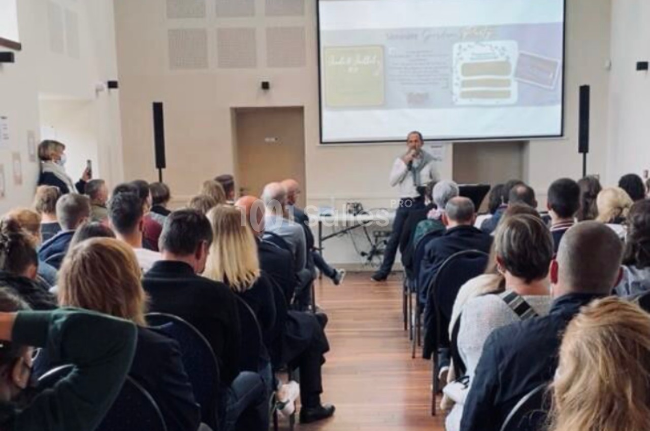 Un conférencier s'adresse à un public assis dans une salle, avec une présentation projetée à l'écran.