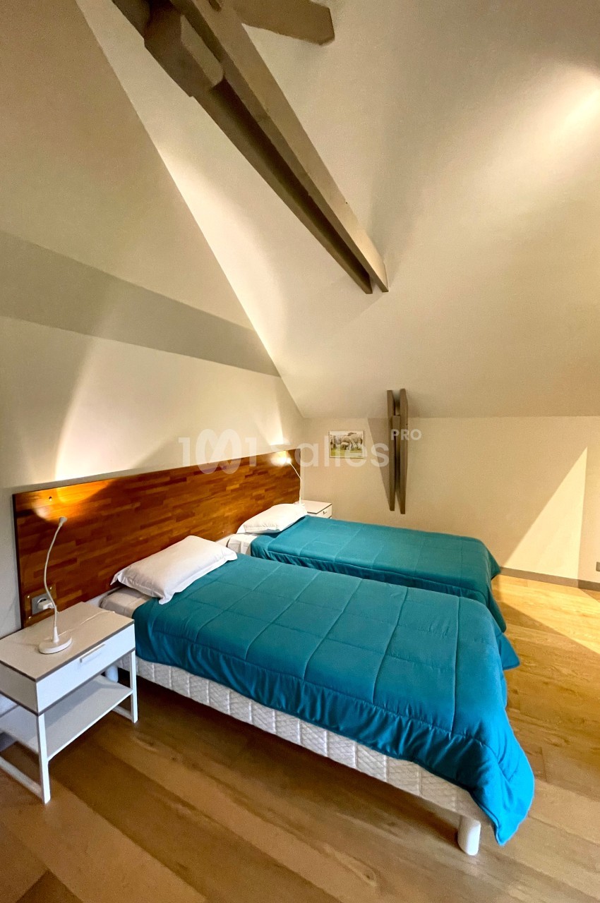 Deux lits simples avec couvre-lits bleus dans une chambre mansardée lumineuse, équipée d'une table de chevet et d'une lampe.