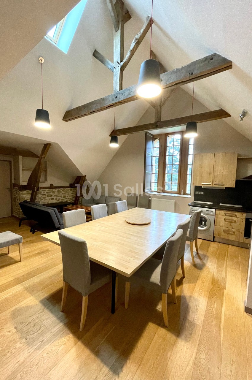 Salle à manger lumineuse avec table en bois, chaises grises, poutres apparentes et cuisine ouverte en arrière-plan.