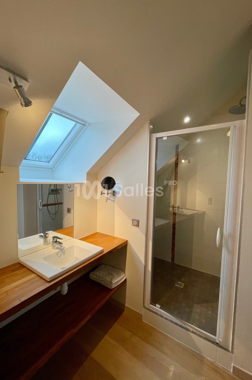 Salle de bain mansardée avec lavabo sur plan en bois, miroir, douche vitrée et fenêtre de toit.