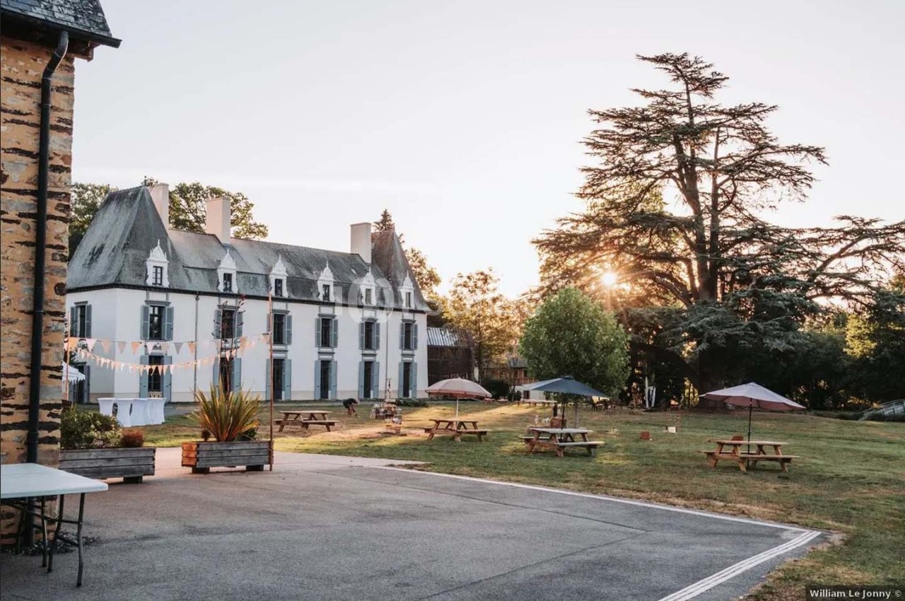 Cour d’un château avec pelouse, tables de pique-nique, parasols et arbres, éclairée par une lumière de fin de journée.