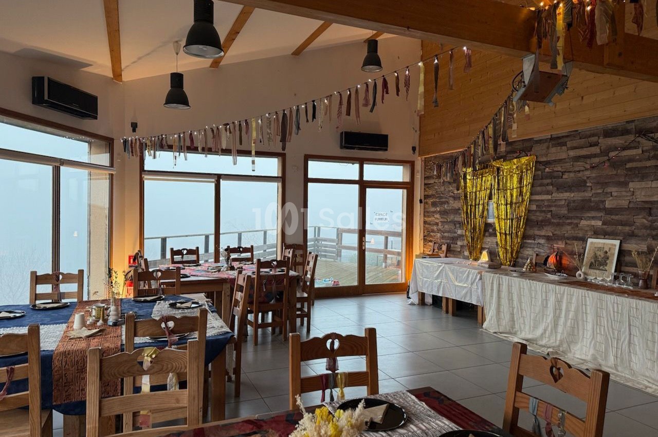 Salle de restaurant lumineuse avec grandes baies vitrées, tables dressées et décorations suspendues.