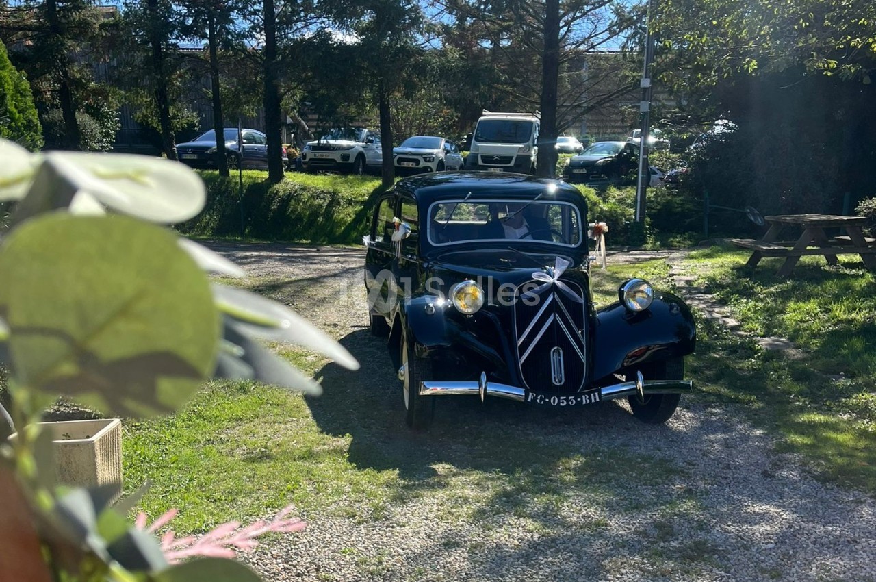Voiture ancienne noire circulant sur un chemin gravillonné entouré de verdure et de véhicules stationnés.