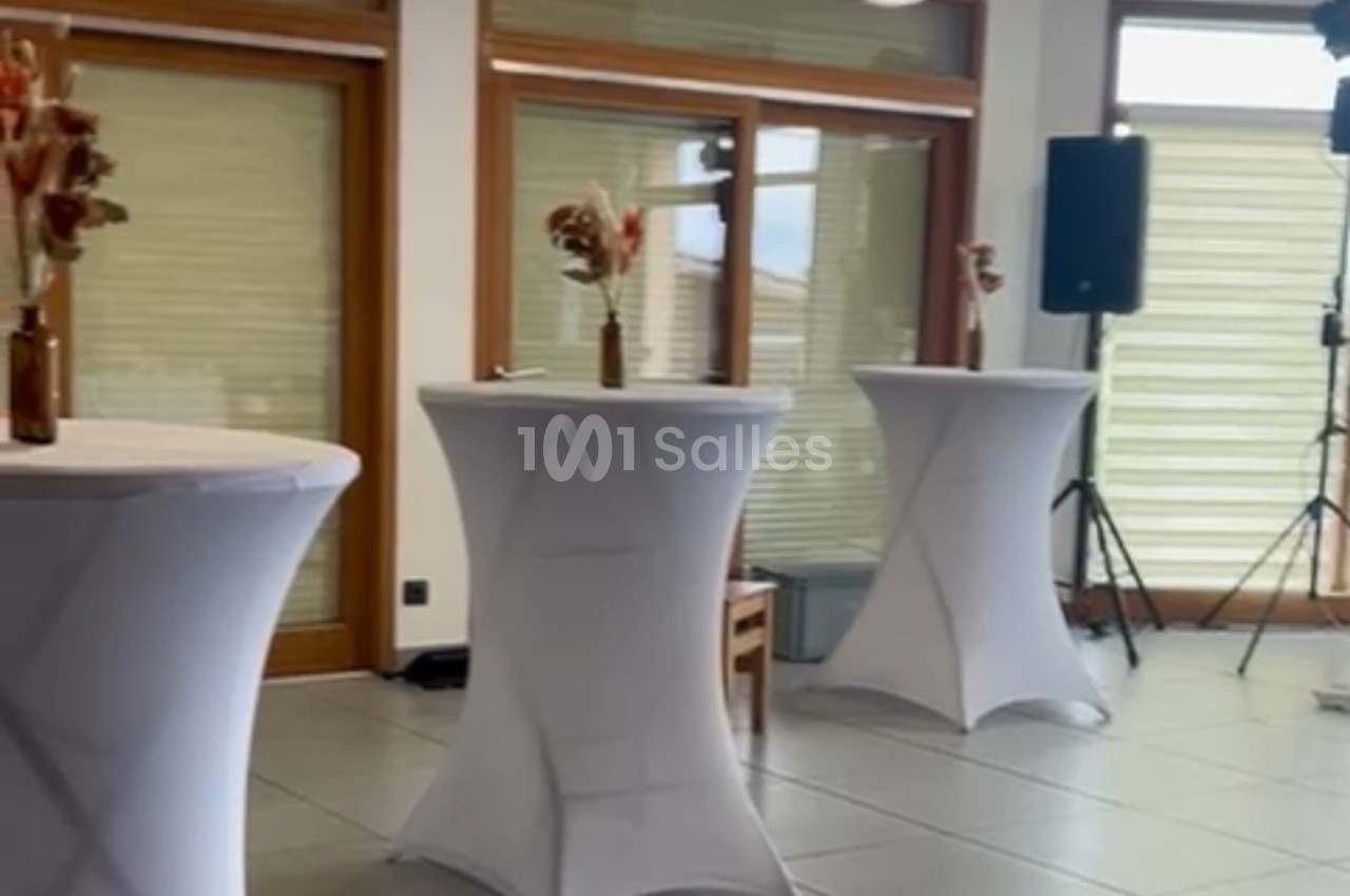 Tables hautes recouvertes de nappes blanches, décorées de bouquets, dans une salle lumineuse avec baies vitrées.