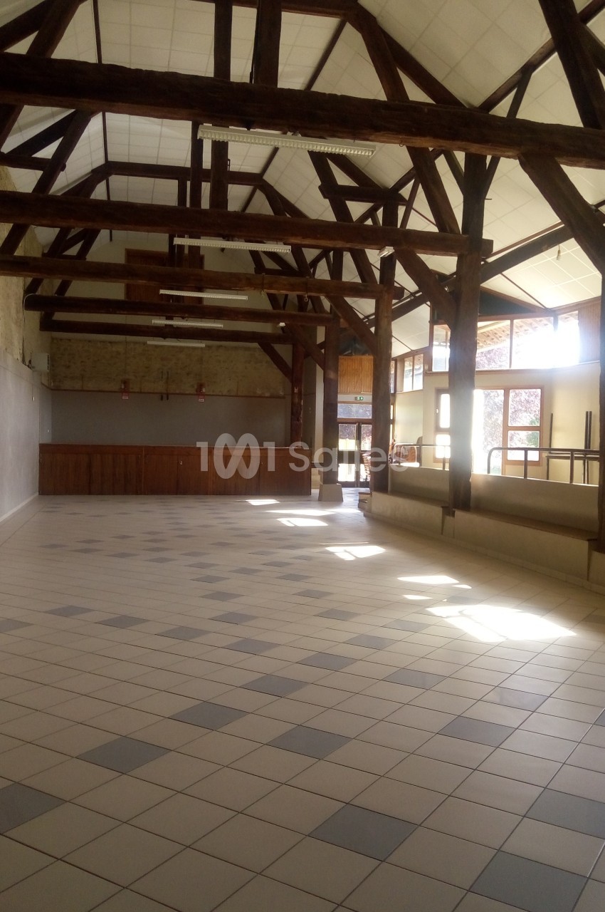 Grande salle lumineuse avec charpente en bois apparente, sol carrelé et murs clairs, éclairée par des fenêtres.