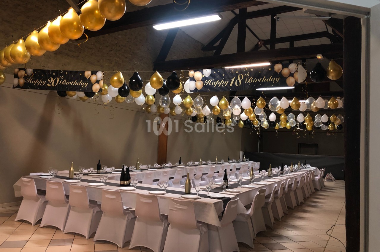 Salle décorée pour une fête avec des tables dressées, des ballons dorés, noirs et blancs suspendus au plafond.