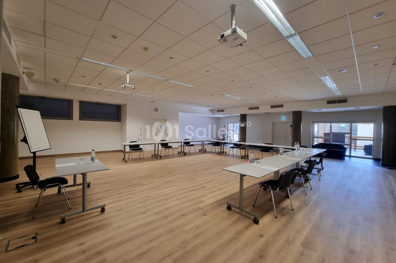 Salle de réunion spacieuse avec tables disposées en U, chaises, tableau blanc et projecteur au plafond.