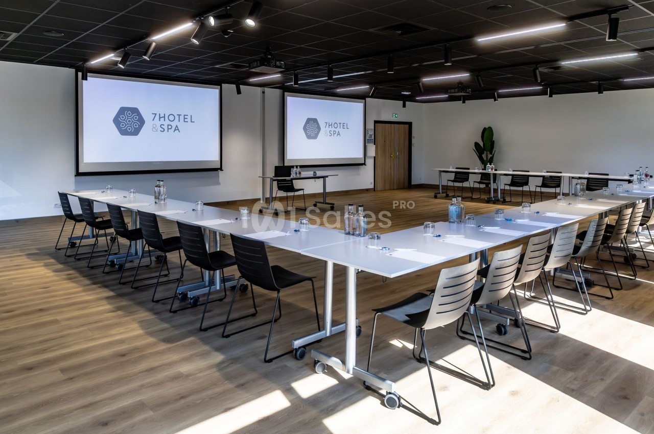 Salle de réunion lumineuse avec tables disposées en U, chaises noires et écrans de projection allumés.