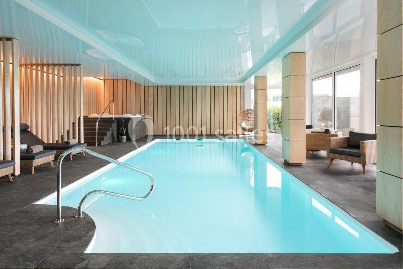 Piscine intérieure lumineuse avec chaises longues, fauteuils et grandes baies vitrées donnant sur l'extérieur.